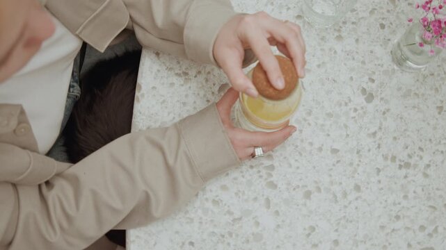Close up of woman in beige holding and placing object into jar on white speckled table. scene includes visible hands clothing details like ring worn by her left hand empty glassware