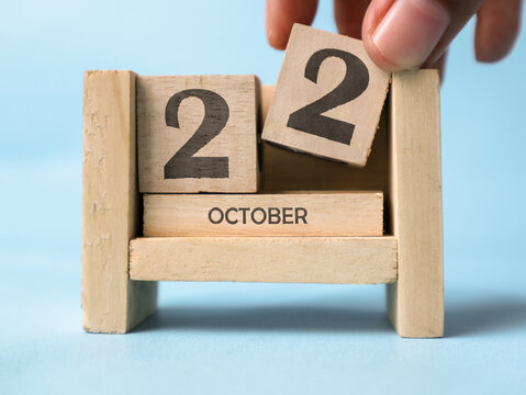 A woman's hand is holding a wooden block with the number 2 written on it on a wooden block calendar for October 22nd on a light blue background.