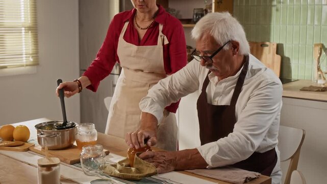 Senior Caucasian man closing homemade jam jar while middle aged Caucasian woman ladling fruit preserve in kitchen. Footage ideal for retirement hobby promotions and organic food advertising