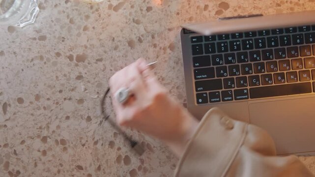 Close up of woman hand interacting with open laptop on speckled stone countertop connecting and disconnecting USB C cable. Two glasses containing water are visible in background. Soft warm