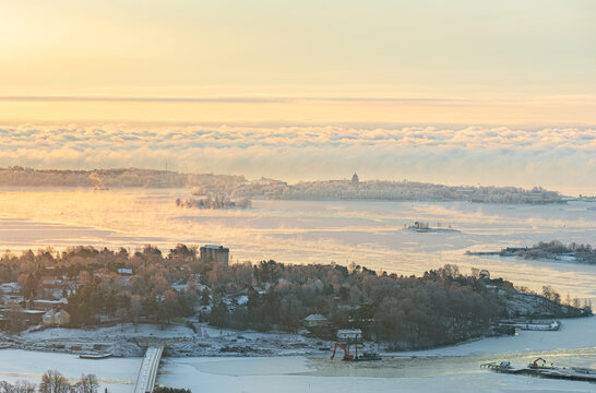 Aerial view of the frozen Helsinki archipelago and coastline covered in snow during a golden sunset, featuring islands, icy waters, and urban buildings in Helsinki, Uusimaa, Finland.