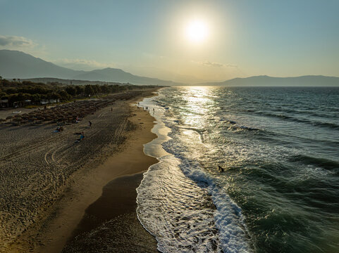 Aerial view of the sandy beach with rows of straw umbrellas, gentle waves rolling onto the shore, and the sun setting over the distant mountains in Chania, Greece.