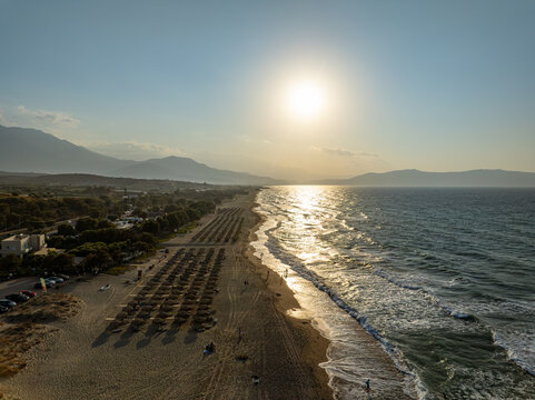 Aerial view of the beach in Chania with rows of sun umbrellas, gentle waves rolling onto the sand, and mountains under a golden sunset sky in Chania, Greece.