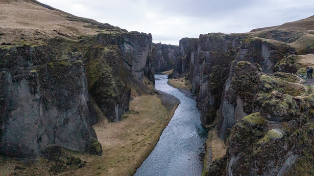 Aerial view of the winding river flowing through the deep rocky canyon of Fjadrargljufur with moss-covered cliffs under an overcast sky in Iceland.