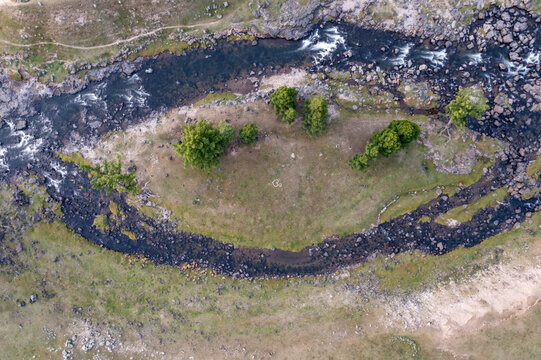 Aerial view of the winding river surrounding a grassy island with scattered trees and rocky banks near the Red Waterfall in Burgaasatayn Hiid, Ovorkhangai, Mongolia.