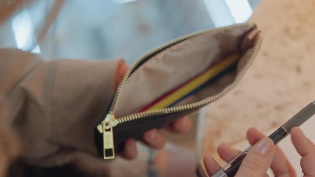 Close up of woman hand holding and examining various pens over open notebook. pencil case with colored pencils is visible in background indicating preparation for writing or drawing. scene