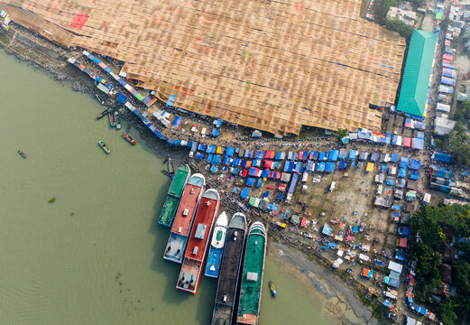 Aerial view of the Charmonai riverbank with passenger ferries docked along the shore and a sprawling market area with colorful stalls in Charmonai, Barisal Division, Bangladesh.