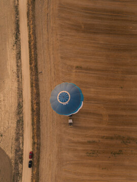 Aerial view of a blue and white hot air balloon hovering above a dry agricultural field next to a dirt road with two parked cars in Gravina in Puglia, Apulia, Italy.