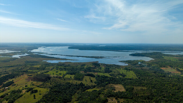 Aerial view of the expansive Parana River winding through lush green forests and agricultural fields under a clear blue sky in Nueva Alborada, Itapua Department, Paraguay.