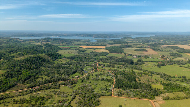 Aerial view of the lush rural landscape featuring rolling green hills, dense forests, winding dirt roads, and a distant river in Nueva Alborada, Itapua Department, Paraguay.