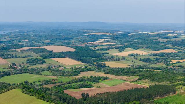 Aerial view of rolling agricultural landscape with patchwork fields, dense green forests, and distant hills under a clear sky in Nueva Alborada, Itapua Department, Paraguay.