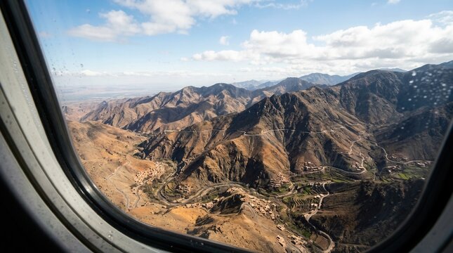 Aerial view of Atlas Mountains from airplane window, showcasing winding roads, terraced fields, and traditional villages in a rugged landscape.