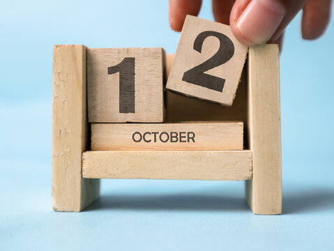 A woman's hand is holding a wooden block with the number 2 written on it on a wooden block calendar for October 12th on a light blue background.