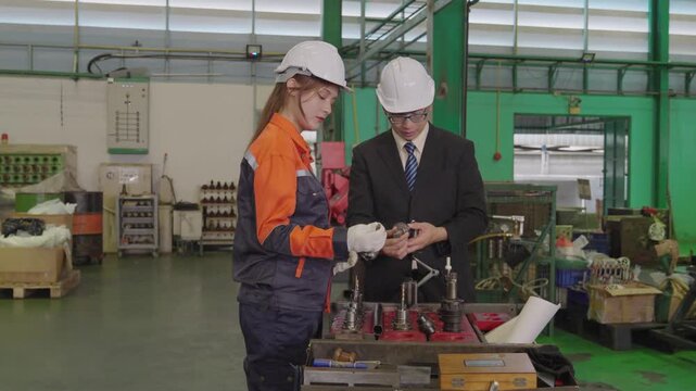 Factory worker and manager wearing hard hats examining machine parts together in industrial workshop, engineer and technician discussing manufacturing equipment, teamwork.