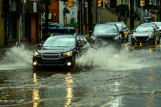 Vehicles drive through a flooded commercial street after a heavy rainstorm, splashing through standing water at dusk