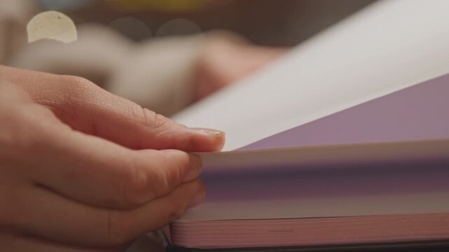 Close up of woman hands turning pages of open notebook with soft lighting. focus remains on hand movement and page interaction showing texture detail in skin closeup. Background is blurred