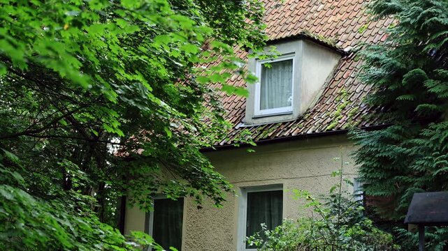 Old house with tiled roof and attic window hidden among green trees. Rustic cottage facade in summer forest. Tranquil nature and vintage architecture concept.