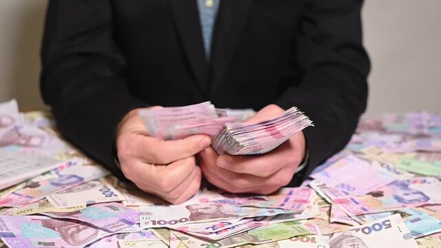 Man counts Ukrainian hryvnia banknotes sitting at desk covered with cash. Successful businessman calculates corporate income, managing financial investments. Male worker holds money stack.