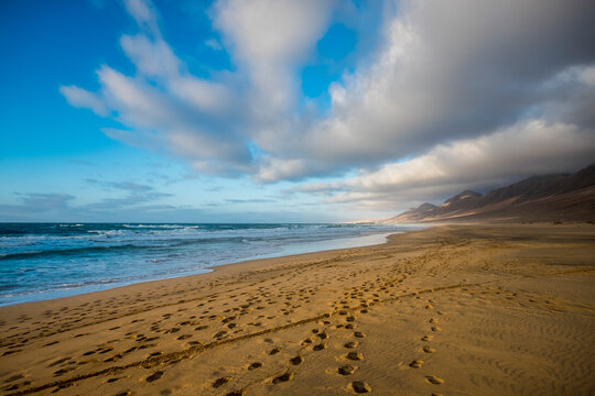 Cofete beach on fuerteventura, canary islands, showing footprints on golden sand, powerful ocean waves breaking, and distant jandia mountain range under a partly cloudy sky