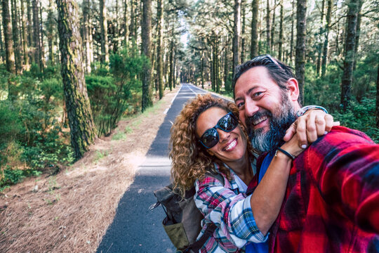 Happy smiling couple hugging and taking a selfie on a sunny forest road, enjoying a hiking trip together, backpacks on, looking at camera, carefree outdoor adventure and love