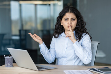 Businesswoman gesturing for silence with a finger on her lips, sitting at her desk with a laptop,...