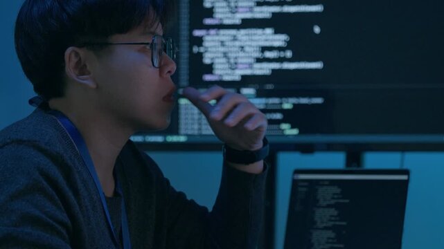 Asian Programmer Thinking While Reviewing Software Code on Multiple Computer Screens at a Workstation in a Dark Blue Technology Office During a Late Night Cybersecurity Project