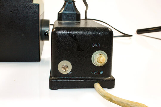 Close-up of a vintage microscope power supply with a 220V switch, control knob, electrical cable and black metal housing on a white background.