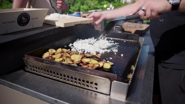 Sliced potatoes turn golden on a hot griddle while a cook adds freshly chopped onions from a wooden board