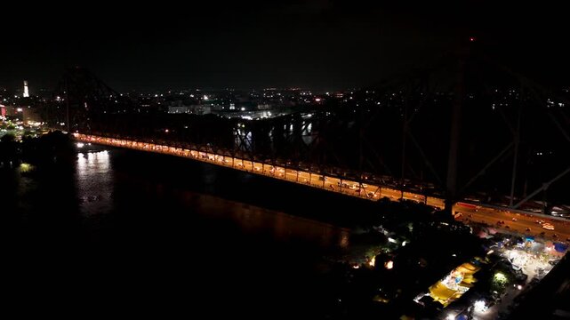 Wide aerial drone arc shot of the illuminated Howrah Bridge at night, with traffic crossing the bridge and the Hooghly River visible below.