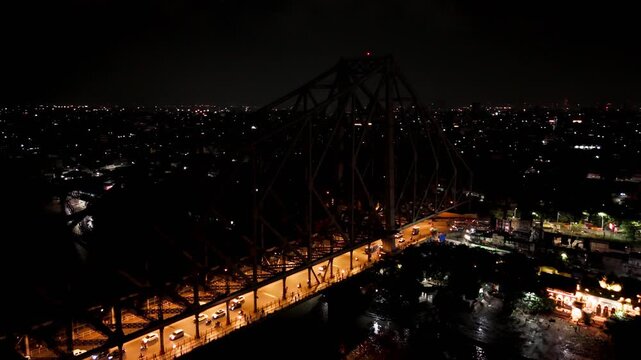 Aerial drone arc shot around illuminated Howrah Bridge at night, showing traffic crossing the bridge with Kolkata city lights in the background