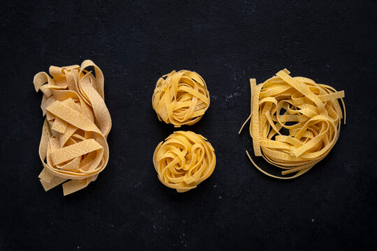 Dry food pasta - carbohydrate Italian traditional meal ingredient. Assortment with raw pappardelle ribbons and tagliatelle nests on black background. Premium gourmet healthy eating