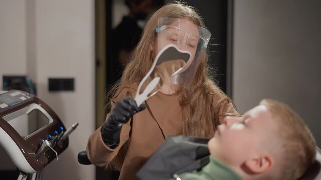 Young woman wearing face shield and brown top demonstrates dental care to boy reclining in orthodontic chair holding up tooth shaped cutouts for explanation. scene takes place indoors