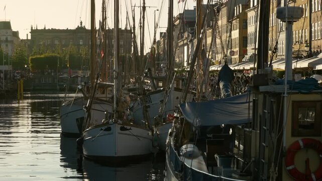 Historic wooden sailboats moor along the Nyhavn canal in Copenhagen. Tourists relax at waterfront cafes along the vibrant promenade. Danish maritime culture highlights European summer vacation.