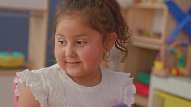 Girl smiling and holding a blue number card with her hand while seated at a pink chair at a toy table inside a daycare building filled with learning toys; playful learning discovery.