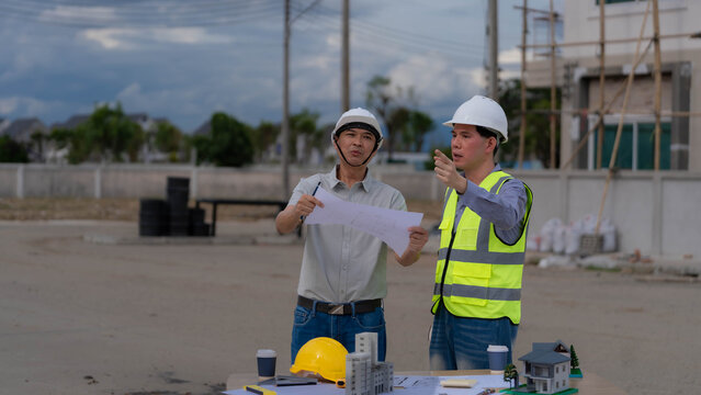 Two Asian Engineers Discussing Blueprint with Architectural Models Outdoor at Construction Site
