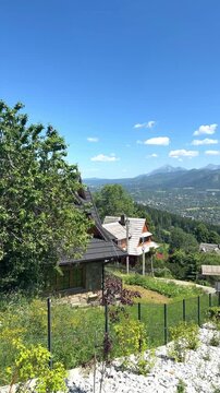 Traditional wooden village nestles on hillside overlooking mountain ridge under blue sky. Rural countryside features lush trees and garden. Scenic resort village offers quiet vacation. vertical