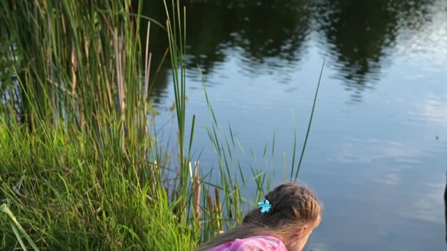 Young girl plays with cattail beside calm lake, holding stem near water. Outdoor play encourages nature discovery, while cattail adds wetland texture and peaceful background for summer recreation.