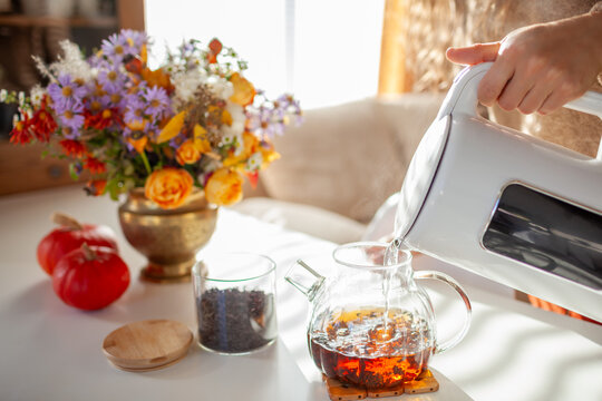 Drinking tea at home decorated for autumn with flowers, pumpkins. Pouring hot water in glass teapot. Cozy fall interior decor 
