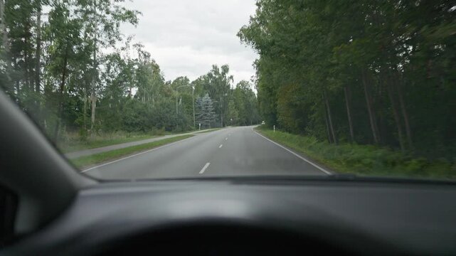 Curving forest road seen from car interior with tall trees, empty lane markings, and overcast sky on quiet rural drive