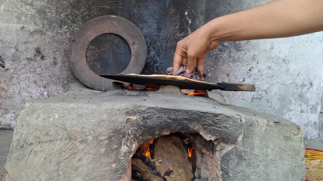 Village woman flipping traditional flatbread roti on hot iron tawa pan over a wood fire clay stove in a rustic kitchen   