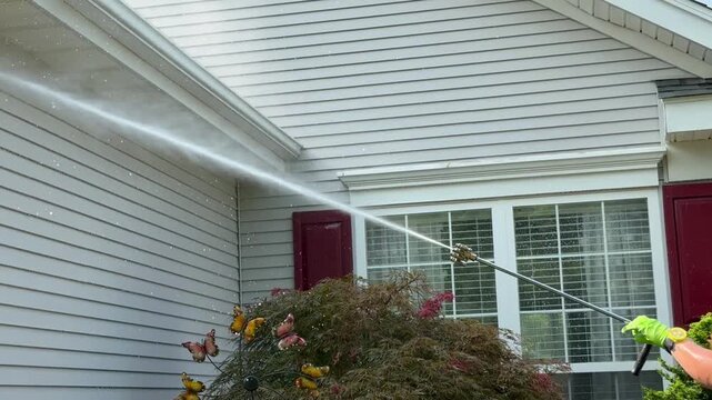 Gloved hand holding a power washer wand spraying water at high pressure to clean the siding and gutters on the side of a gray house