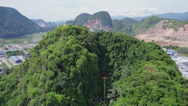 Drone flies above a densely forested mountain revealing a monastery staircase and Buddhist stupa with towering limestone karsts and developed valley landscape beyond.