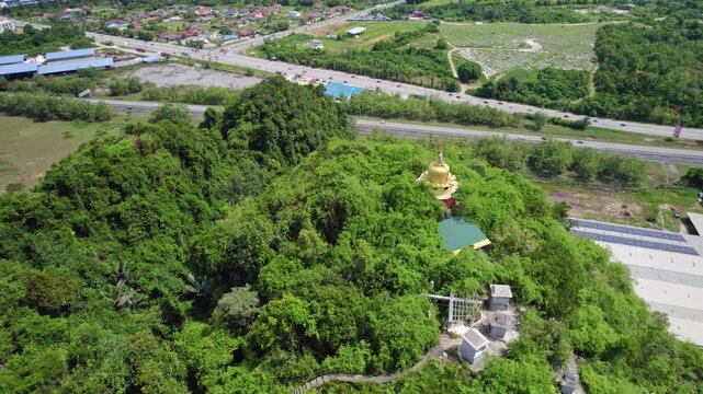 Drone footage passing above a forested hilltop Buddhist monument overlooking modern transportation infrastructure and surrounding development in Malaysia.