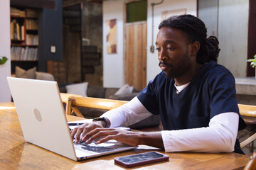 Obraz premium African man wearing scrubs working on laptop at wooden table at home, smartphone nearby, copy space