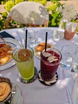 Two colorful cold drinks served in tall glasses on a white table at an outdoor cafe terrace, garnished with dried lime slice and coconut chips on a bright sunny summer day.