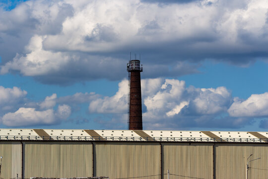 Tall brick industrial smokestack rising against a vibrant blue sky filled with fluffy white clouds, casting a stark silhouette above a modern metal warehouse roof facility