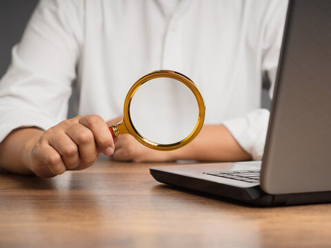 Person holding magnifying glass beside laptop on desk