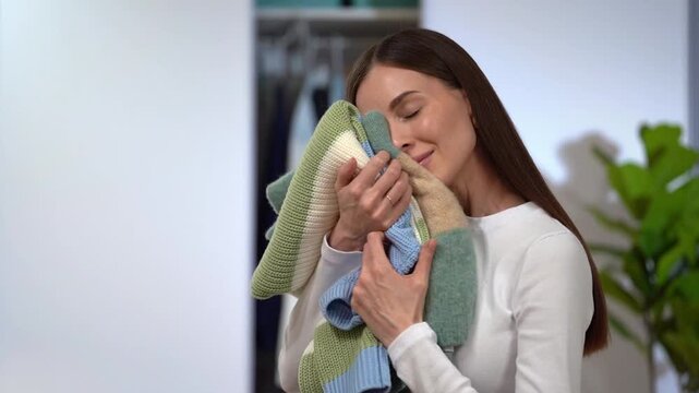 Smiling woman presses soft sweaters to cheek and strokes knitwear by hand on home wardrobe background. Concept of aroma fabric softener, laundry care, garment longevity and eco lifestyle
