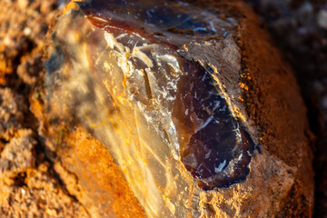 Fototapeta premium Raw translucent chalcedony stone with smooth glassy texture embedded in rough sandy quarry soil, captured in natural bright golden hour sunlight showcasing unique geological mineral patterns
