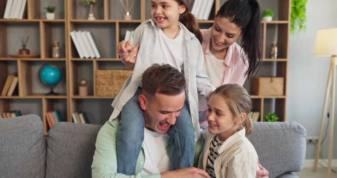 Family parents children playing hugging together at home. Cheerful parents interact with two kids on sofa, sharing laughs, bonding and fun in a bright living room. Love and bonding at home.